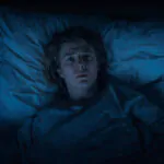 person lying awake in bed at night looking worried at bedside clock showing 3 AM in dimly lit bedroom
